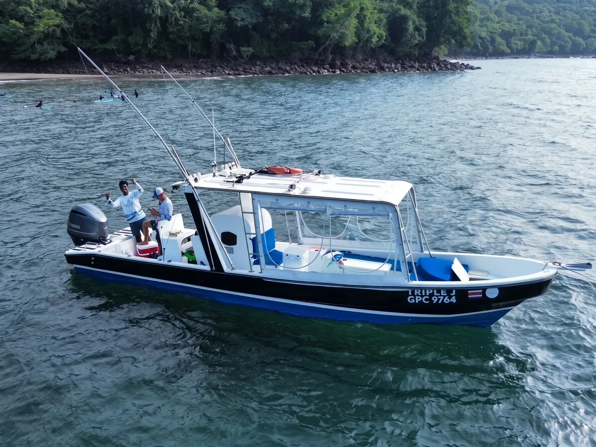 A fishing panga waits in calm water off a wooded shore, two people on its deck, one waving