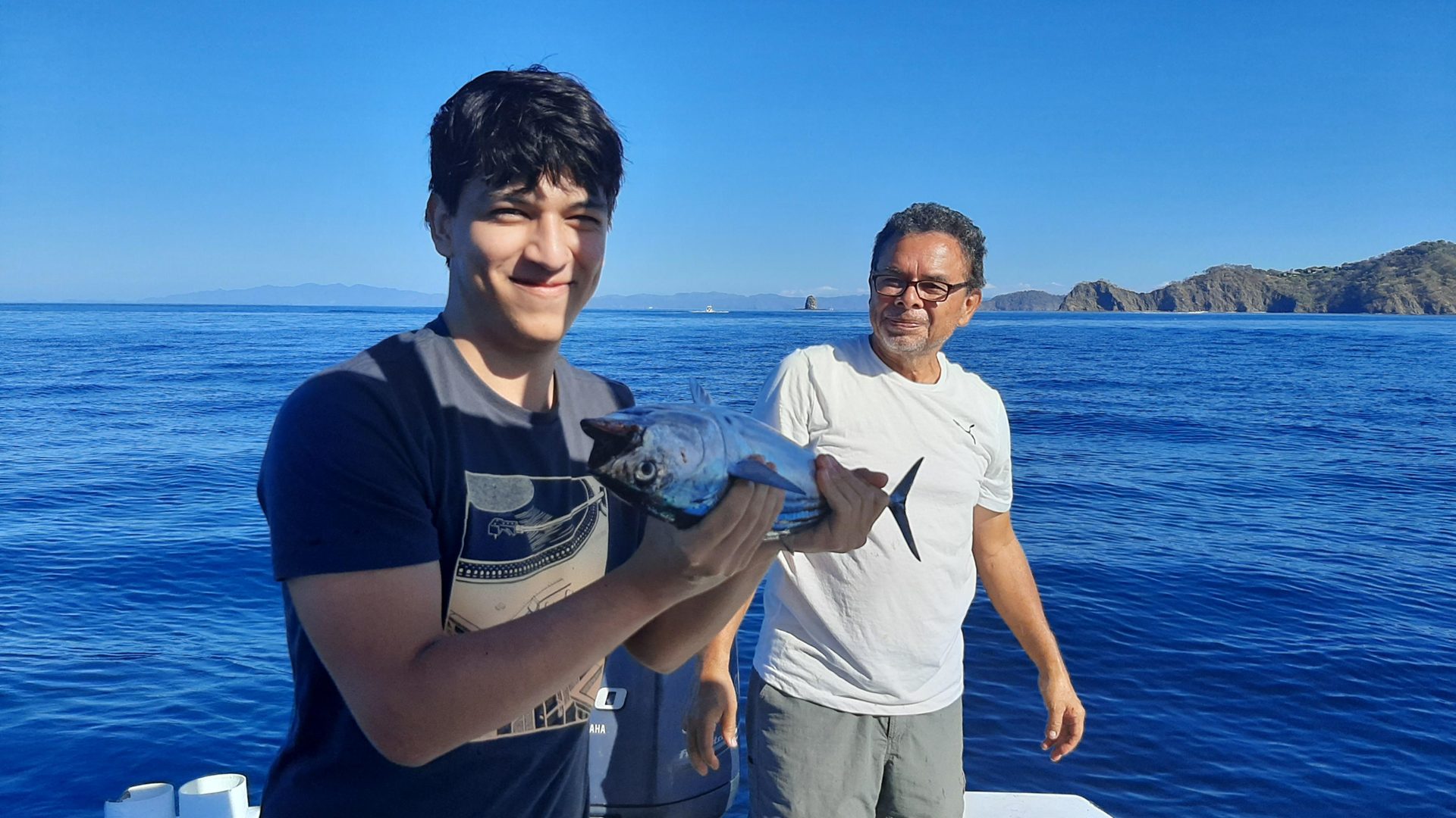 A young angler holds a small tuna on a fishing boat in the Gulf of Papagayo, an older crew member behind him