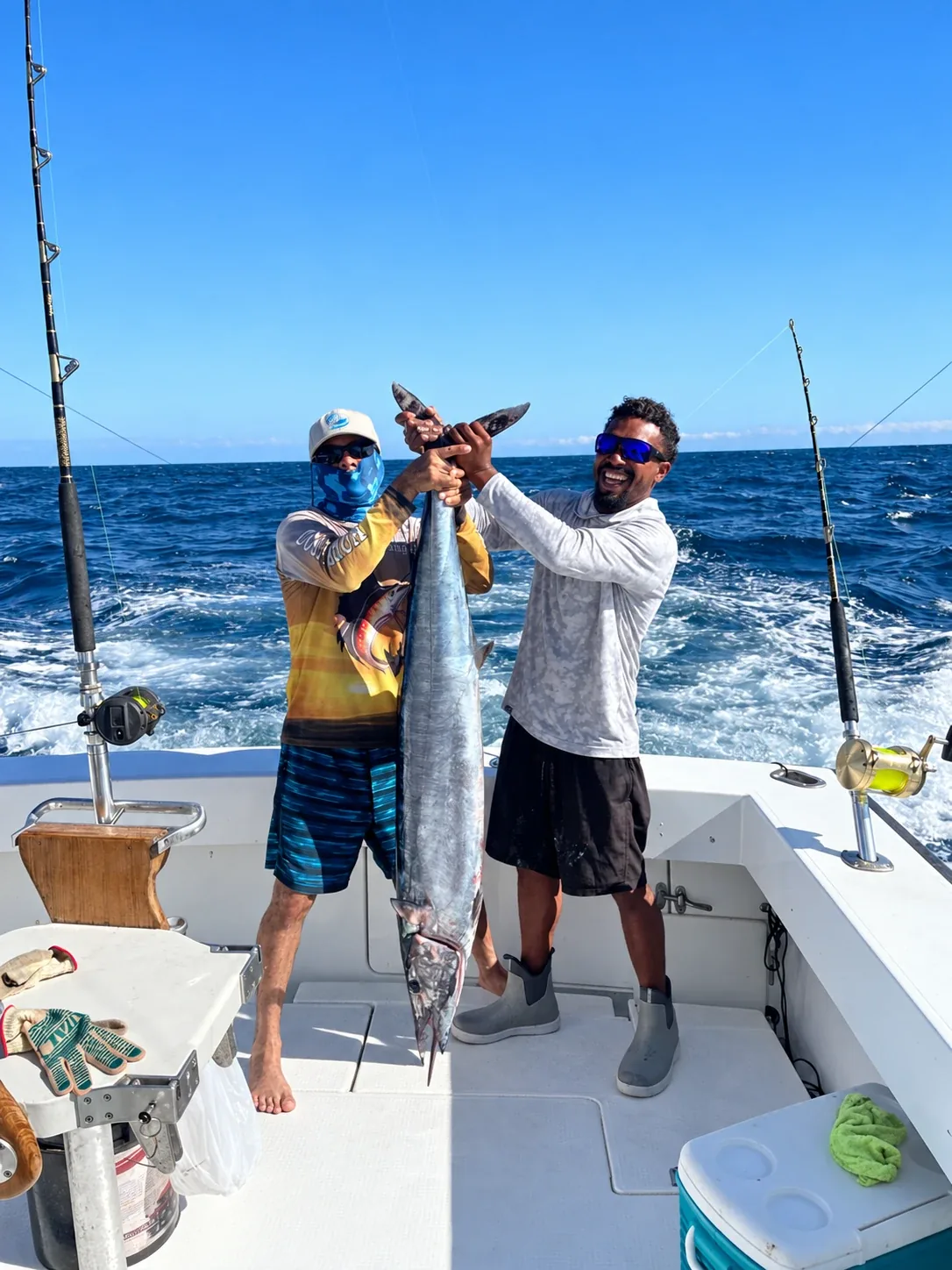 Two anglers hold up a wahoo by the tail on the back deck of a sport fishing boat, with open ocean behind them.